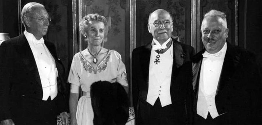Four older adults, three men in tuxedos and one woman in a formal dress, stand together and pose for a black-and-white photograph at a formal event.