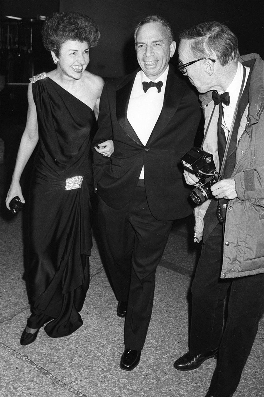 A woman in an elegant black dress and a man in a tuxedo smile, arm in arm, while another man in glasses and a coat holds a camera and laughs with them.