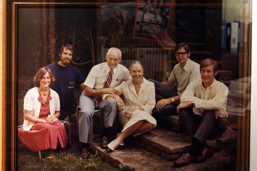Six adults sit and pose outdoors for a group photo, some on stone steps, with greenery in the background. They are dressed in semi-formal clothing and smiling at the camera.