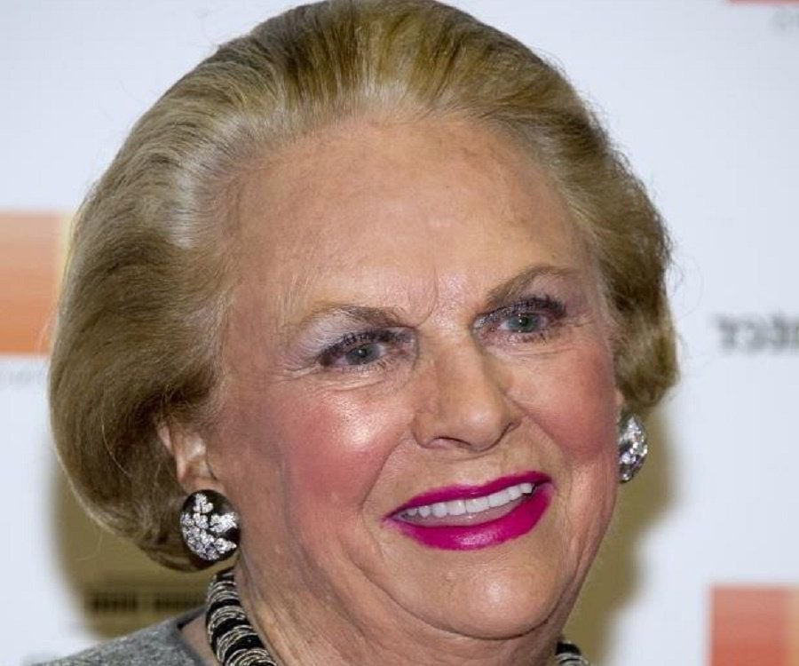 An older woman with styled light brown hair, bright pink lipstick, and rhinestone earrings smiles in front of a blurred background with orange and white colors.
