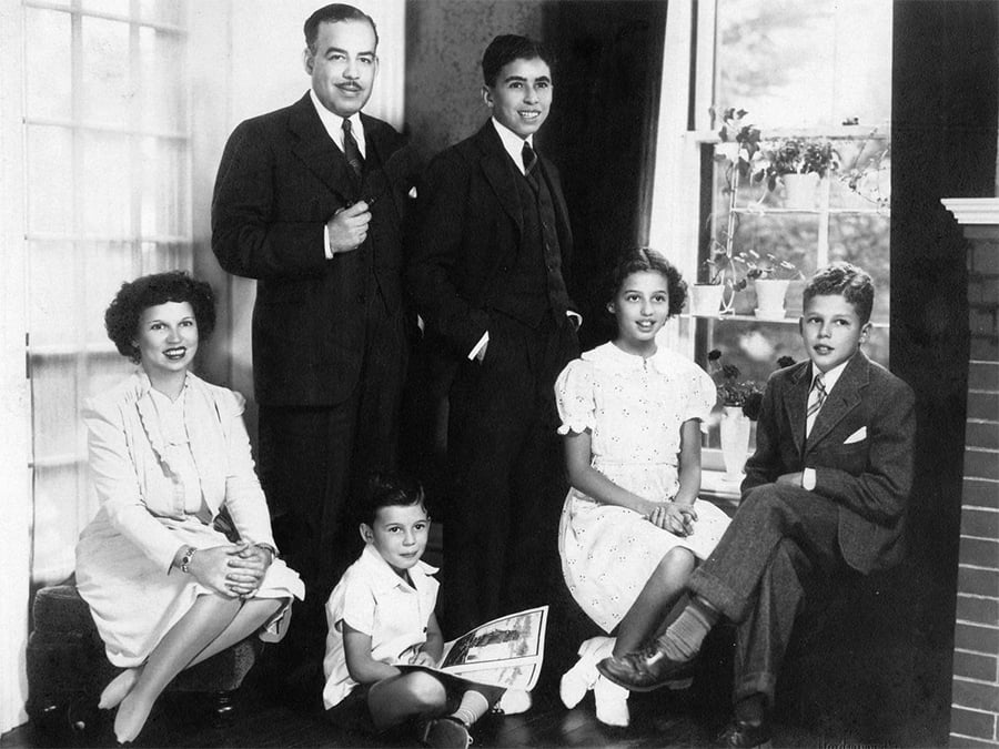 A family of six poses indoors. Two men and a boy stand in suits, while a woman in a dress and two children sit, one holding a book. Sunlight from a window brightens the room with potted plants.