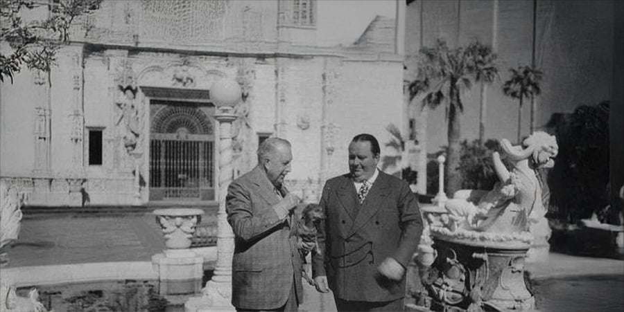 Two men in suits stand by a decorative fountain with statues, in front of an ornate building façade with columns and sculptures. One man holds a small dog. Palm trees are visible in the background.