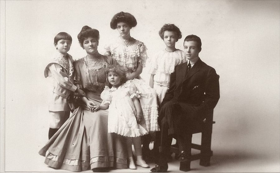 A vintage black-and-white photo of a woman sitting with four children, all dressed in formal, old-fashioned clothing, posing for a family portrait against a plain background.