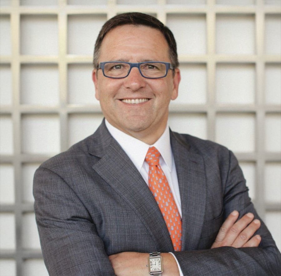 A man in a gray suit, orange patterned tie, and glasses stands smiling with arms crossed in front of a grid-patterned white background.