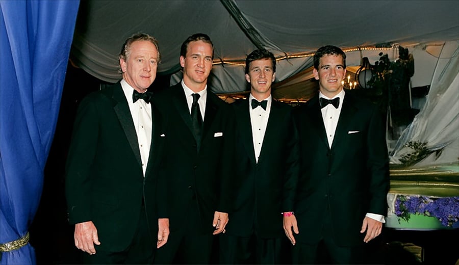 Four men in black tuxedos stand together and smile for a photo at an indoor formal event with draped fabric and blue curtains in the background.