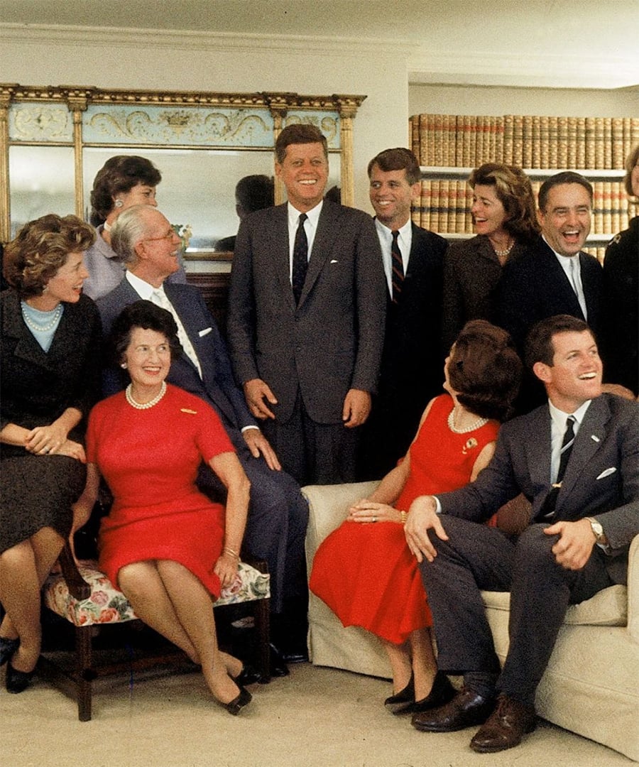A group of well-dressed men and women smile and pose together indoors, with bookshelves and a large mirror in the background.