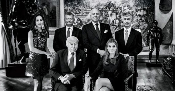 Six well-dressed adults, four standing and two seated, pose in an elegant room decorated with artwork and sculptures. The image is in black and white.