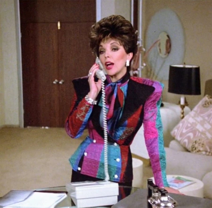 A woman in a colorful, bold 1980s blazer stands in an office, talking on a corded phone. She has styled dark hair and is wearing jewelry. There’s a sofa, lamp, and desk in the background.