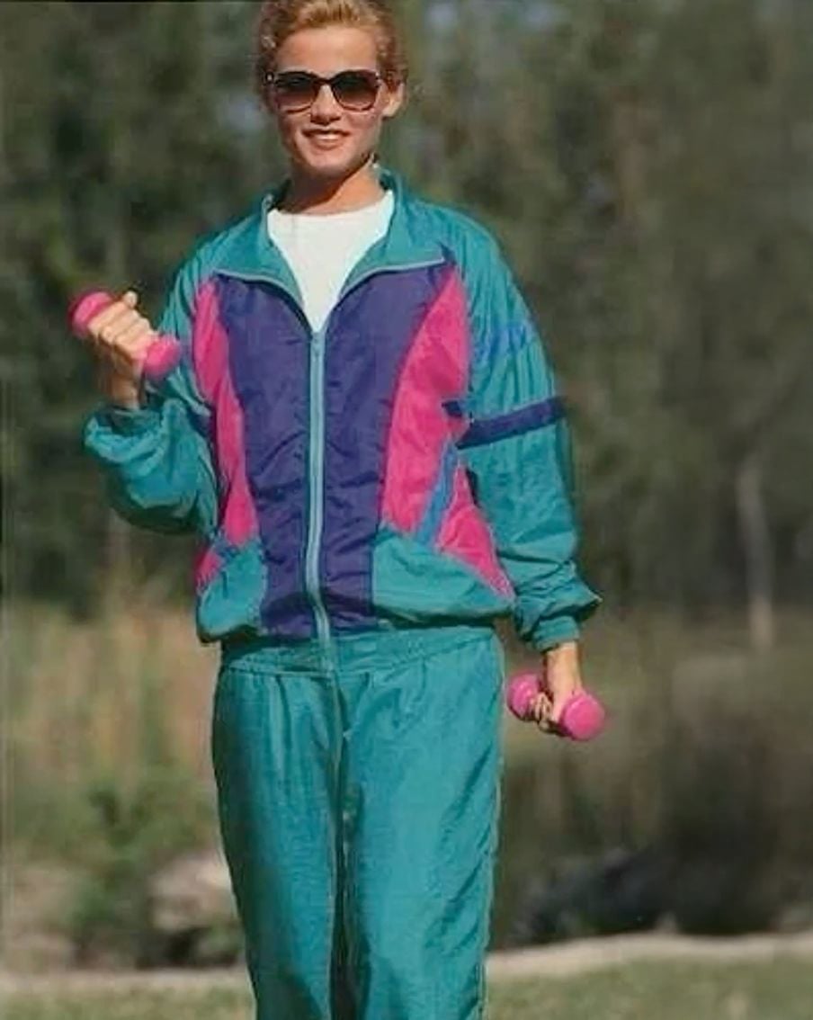 A person wearing a teal and purple 1980s-style tracksuit holds pink dumbbells outdoors, smiling and wearing sunglasses.