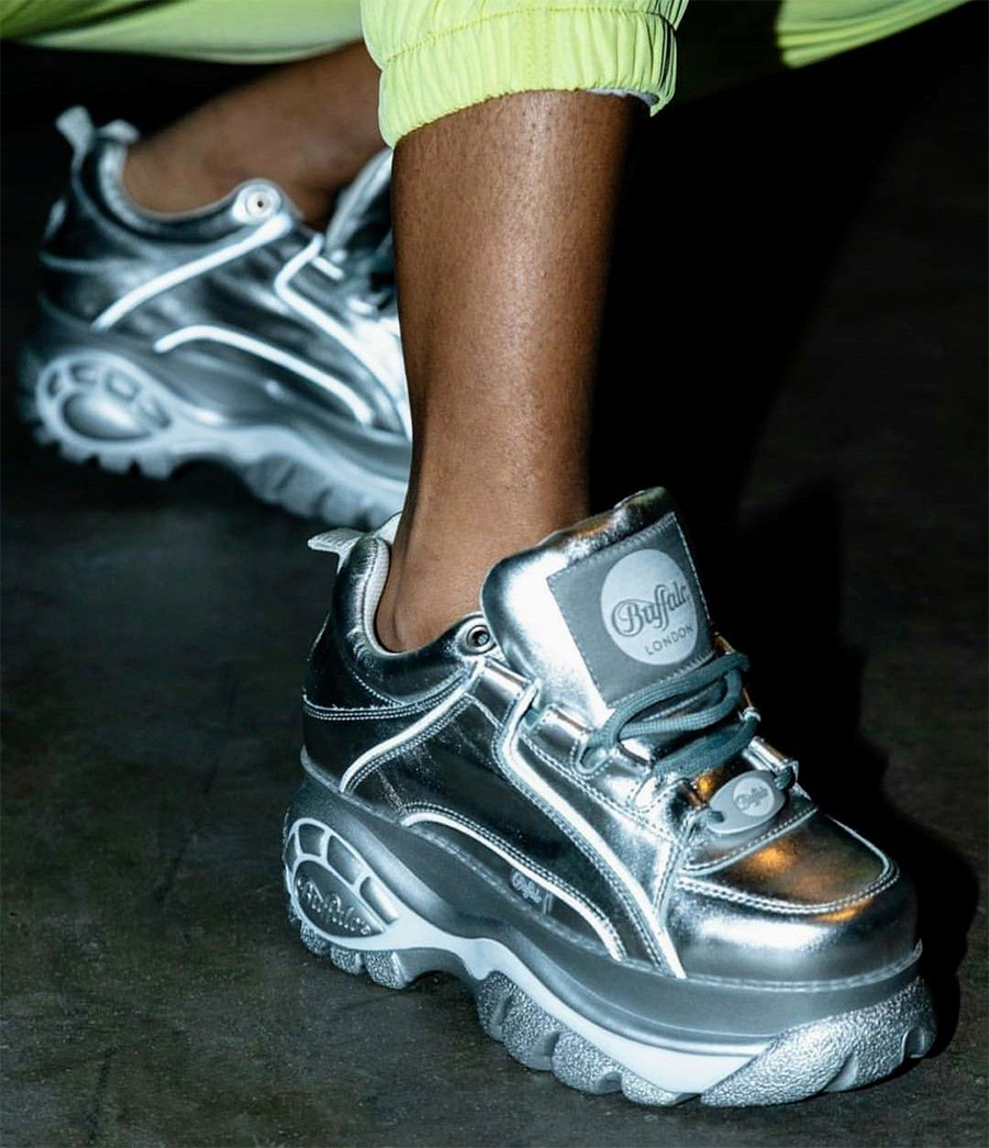 A person wearing metallic silver Buffalo London platform sneakers and yellow-green pants stands on a dark floor.