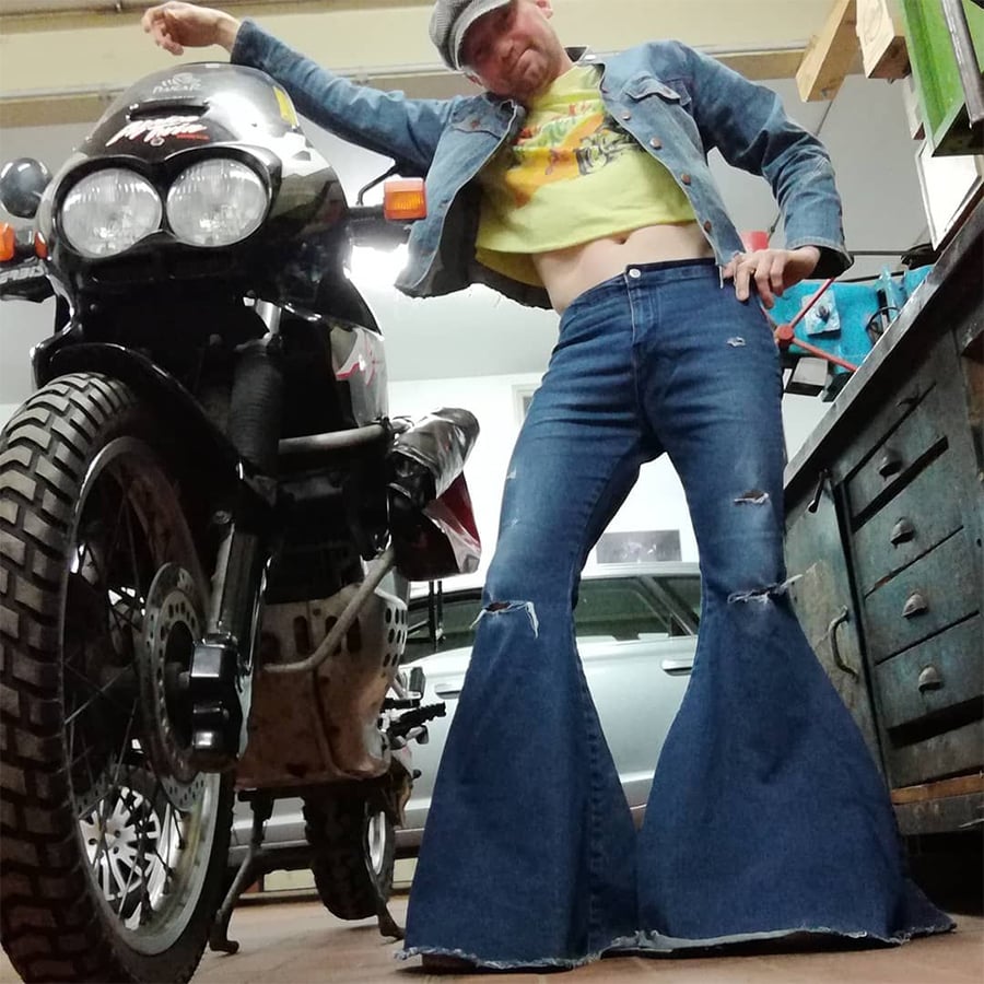 Person posing next to a motorcycle in a garage, wearing a denim jacket, yellow crop top, and exaggeratedly wide bell-bottom jeans, with one arm resting on the bike and the other on their hip.