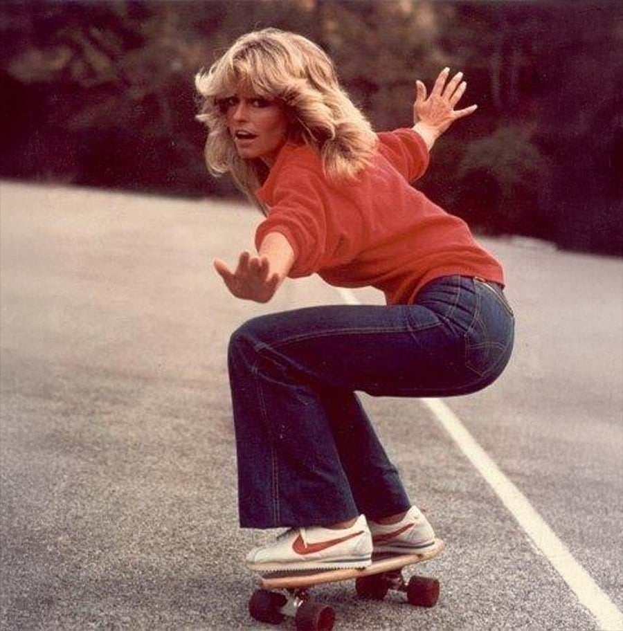 A woman with feathered blonde hair wearing a red sweater, blue jeans, and white sneakers rides a skateboard on a paved outdoor path, balancing with her arms outstretched.