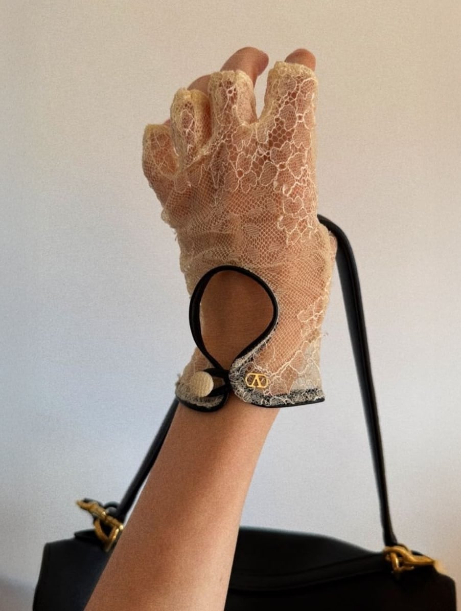 A hand wearing a sheer lace glove with black trim and gold accents holds the handle of a black handbag against a plain background.