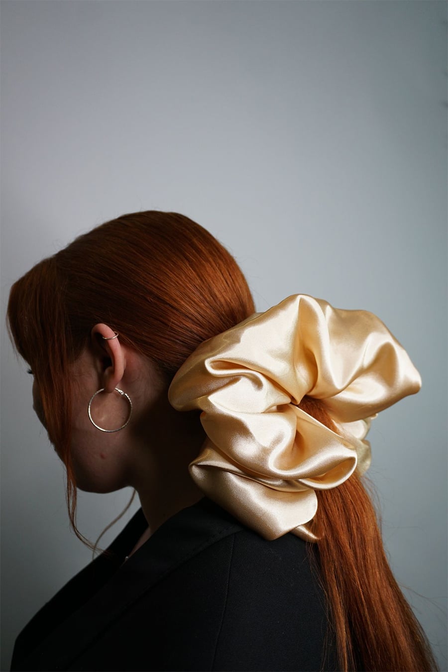 A person with long red hair wears a large, shiny cream-colored scrunchie and a hoop earring, facing away from the camera against a plain background.