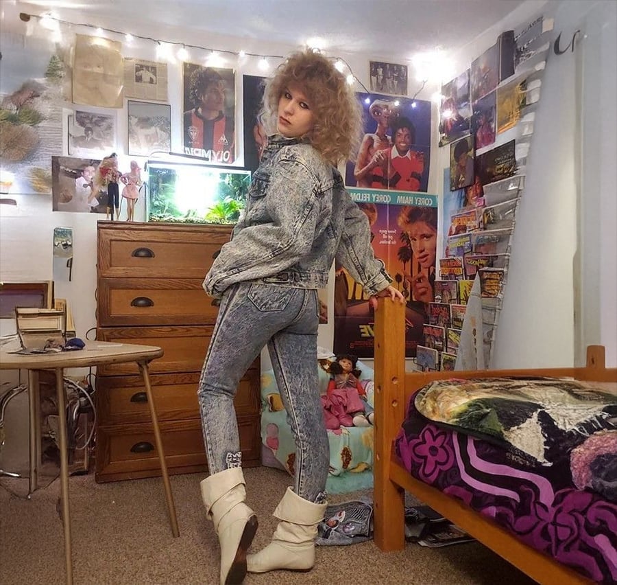 A person with curly hair wears acid-wash denim, white boots, and poses in a bedroom decorated with posters, dolls, string lights, and colorful bedding. The room has a retro, 1980s vibe.
