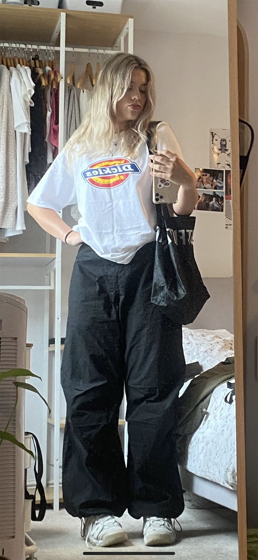 A young woman stands in front of a mirror, wearing a white Dickies T-shirt, black wide-leg pants, and white sneakers. She holds a black tote bag and a phone, with clothing and posters visible in the background.