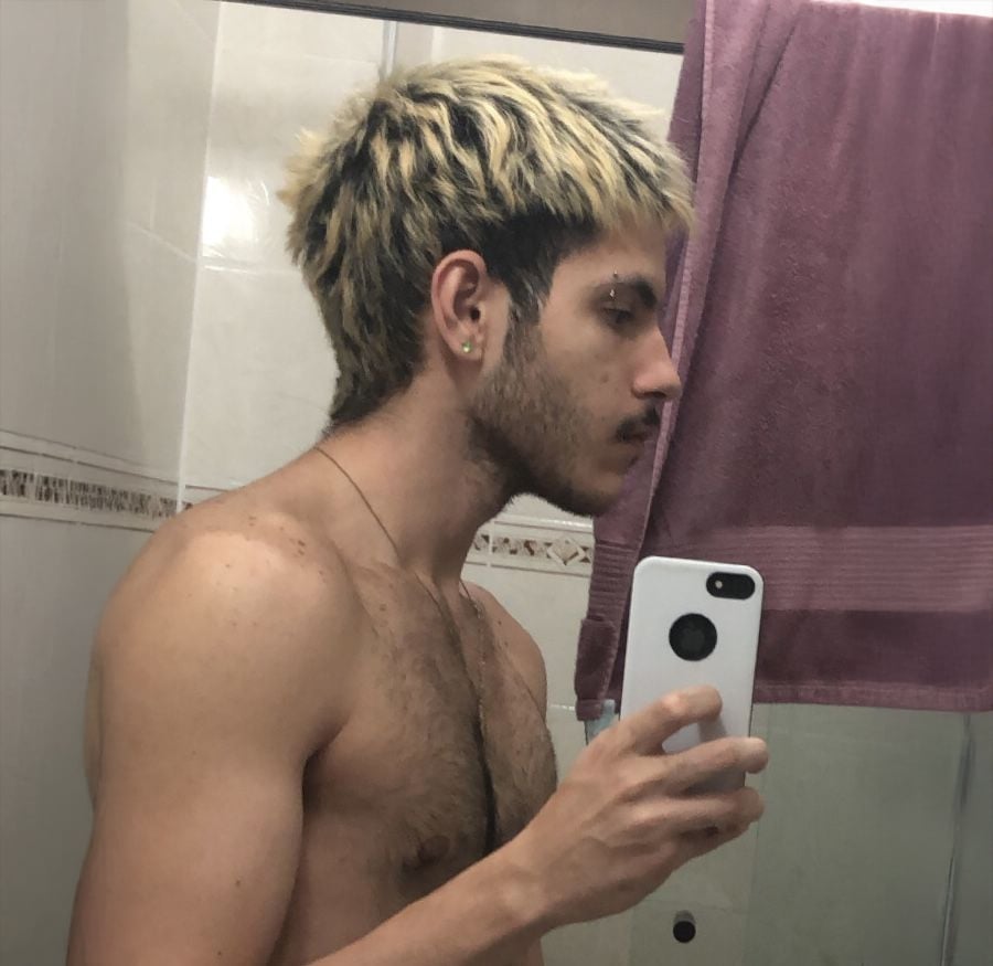A shirtless person with short, blond and dark hair takes a mirror selfie in a bathroom, holding a white phone. A mauve towel hangs on the wall behind them.