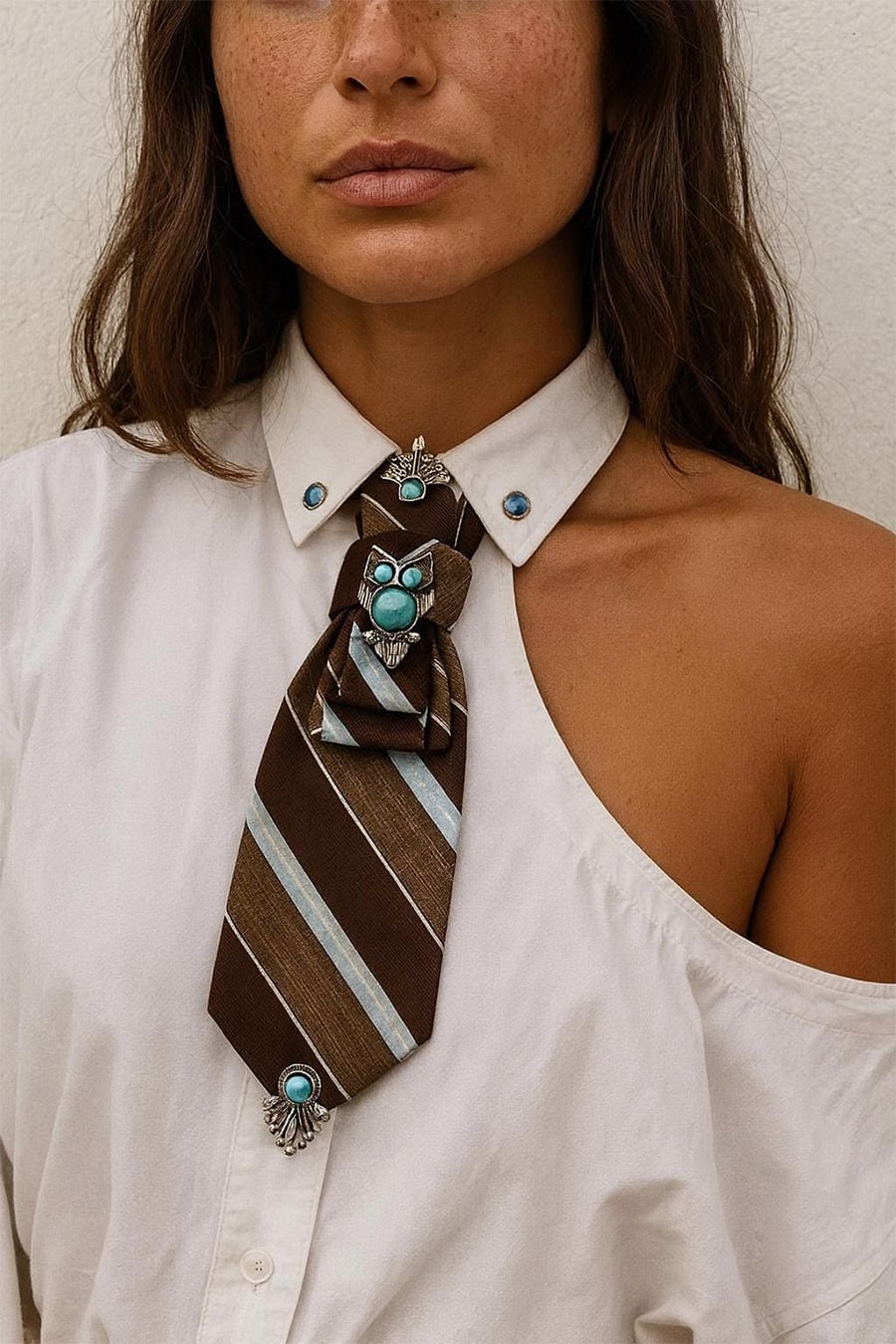 A woman wears a white shirt with one shoulder exposed and a brown striped tie adorned with turquoise jewelry.