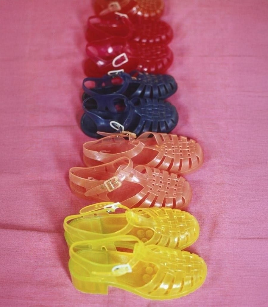 Five pairs of plastic jelly sandals in yellow, orange, blue, red, and maroon are neatly lined up on a pink fabric background.