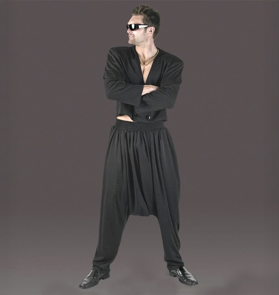 Man wearing sunglasses, a black open jacket, and loose, baggy black pants stands with arms crossed and legs apart against a plain gray background.