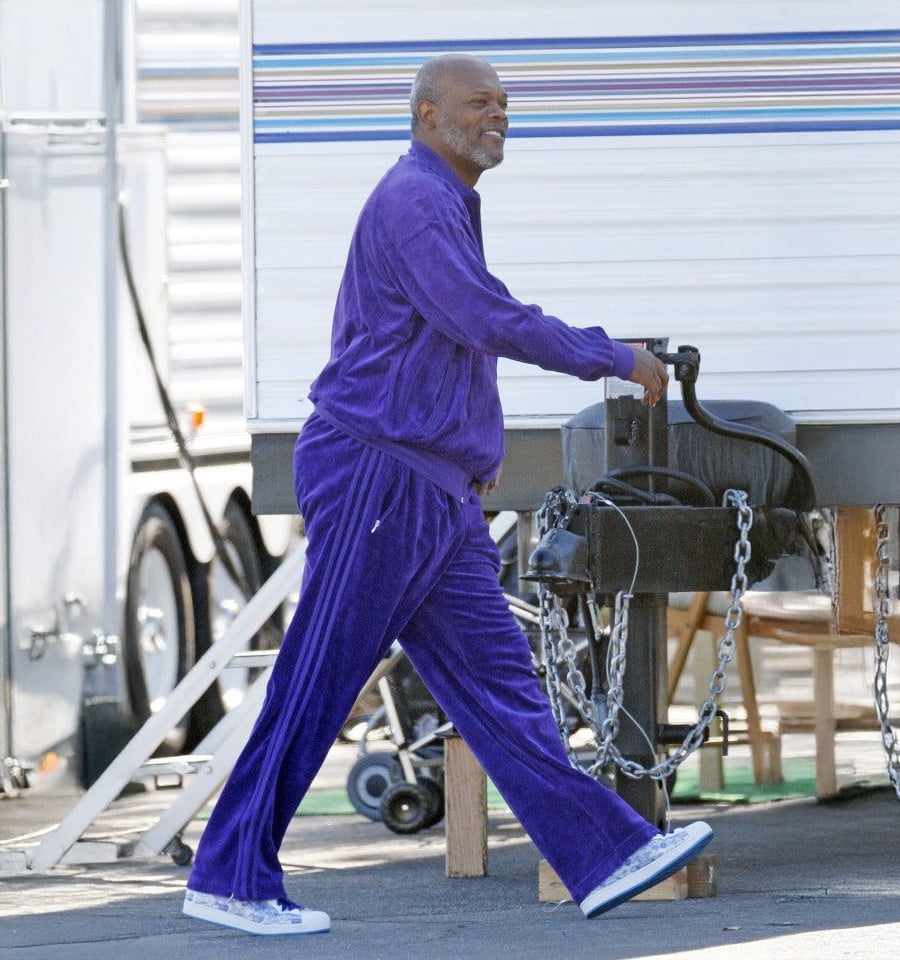 A man in a bright purple tracksuit and white sneakers walks outdoors, smiling, beside a trailer and some equipment.