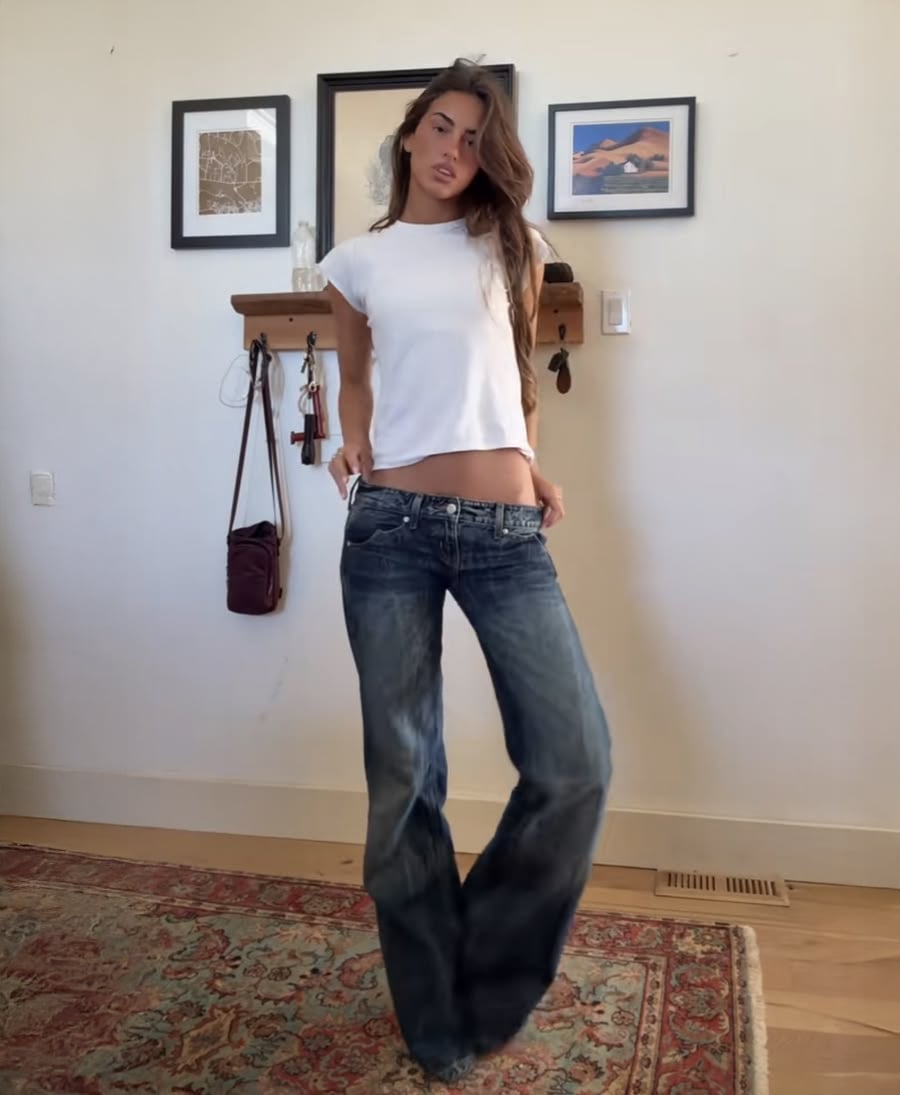 A woman wearing a white t-shirt and blue flared jeans stands indoors on a patterned rug, with wall art, a shelf, and a small bag in the background.