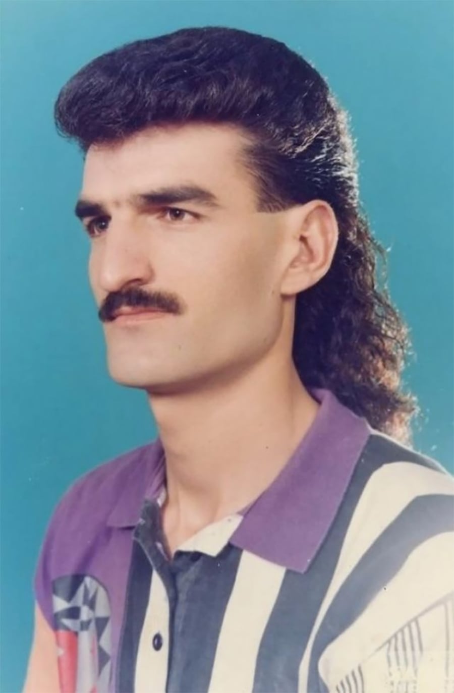 A man with a dark, curly mullet hairstyle and mustache poses for a studio portrait against a blue background, wearing a collared shirt with bold black, white, and purple stripes.