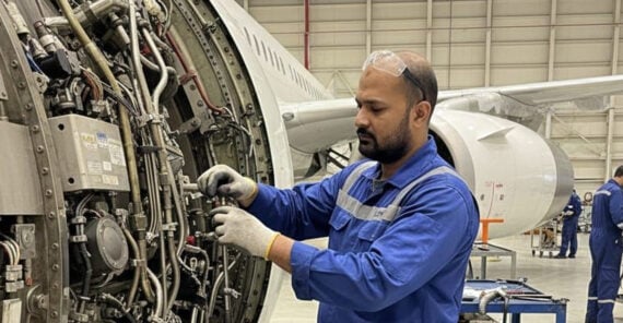 A man in blue coveralls and safety glasses works on an aircraft engine inside a hangar. The engine’s complex wiring and parts are visible, and an airplane is seen in the background.