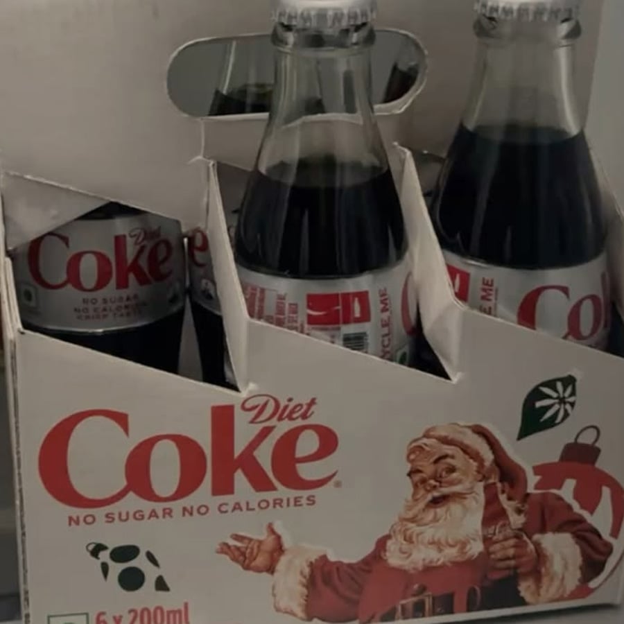 Six-pack of Diet Coke glass bottles in a white festive box featuring a vintage-style Santa Claus illustration. "No sugar, no calories" text is on the packaging, and bottles are partially visible in the box.
