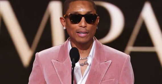 A person in a pink velvet suit and sunglasses speaks into a microphone on stage, wearing earrings and a necklace, with blurred large text in the background.