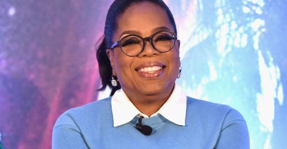 A woman wearing glasses and a blue sweater with a white collar smiles while sitting in front of a colorful, blurred background.