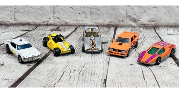 Five toy cars in various colors and styles are lined up on a wooden surface, each facing forward. The cars are white, yellow, silver, orange, and orange with purple and pink details.