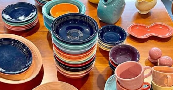 A variety of colorful ceramic dishes, bowls, plates, cups, and pitchers are neatly stacked and arranged on a wooden table.