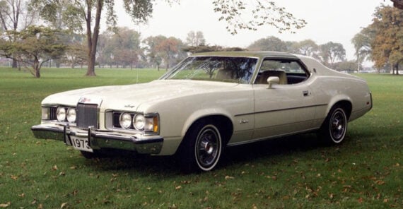 A vintage white 1973 Mercury Cougar parked on grass in a park, surrounded by trees with early autumn foliage in the background.