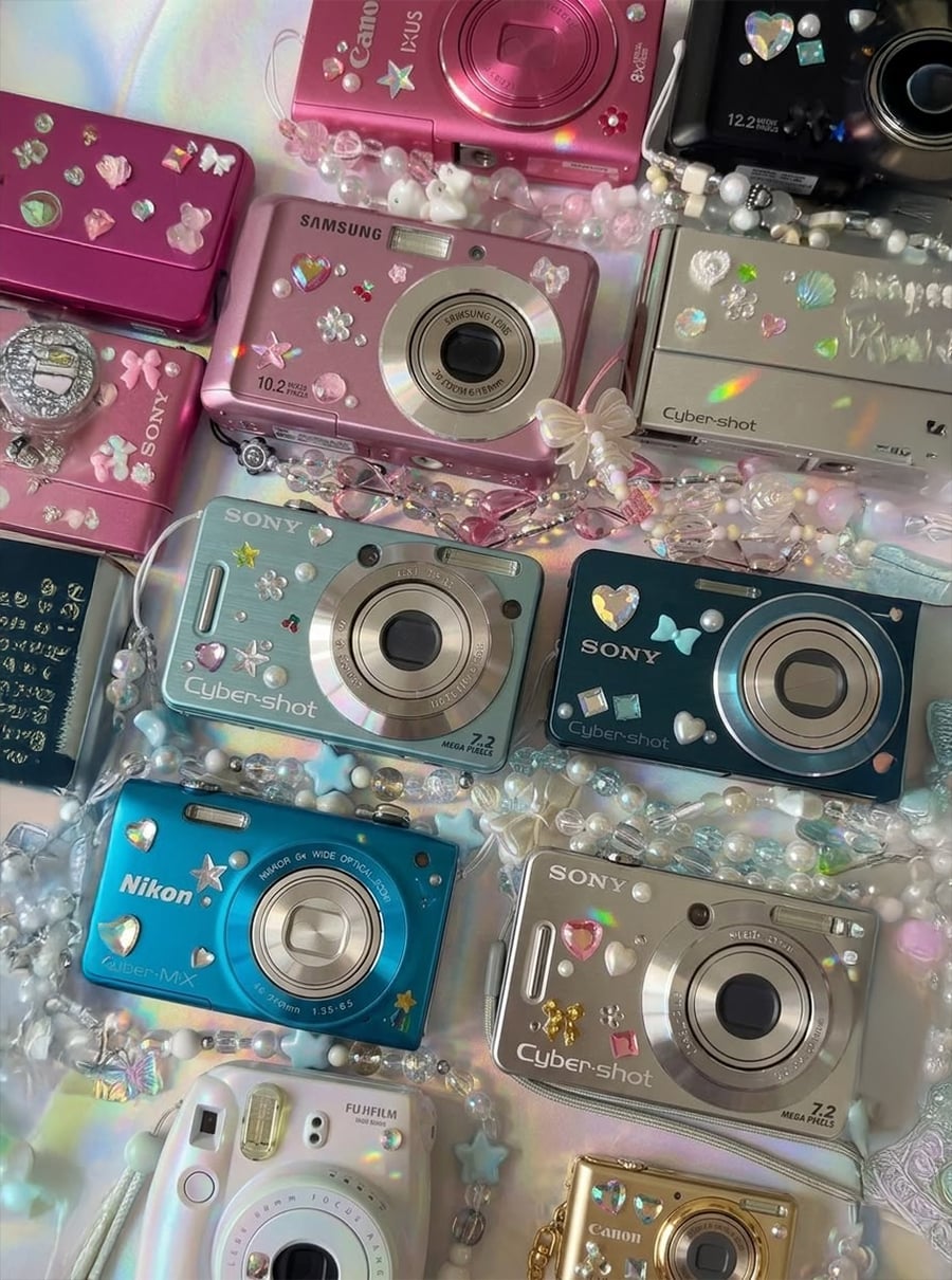 Several colorful digital cameras, mostly Sony Cyber-shot models, are decorated with stickers, pearls, and charms. Cameras in shades of pink, blue, silver, and gold are arranged on a pastel, sparkly surface with beads.