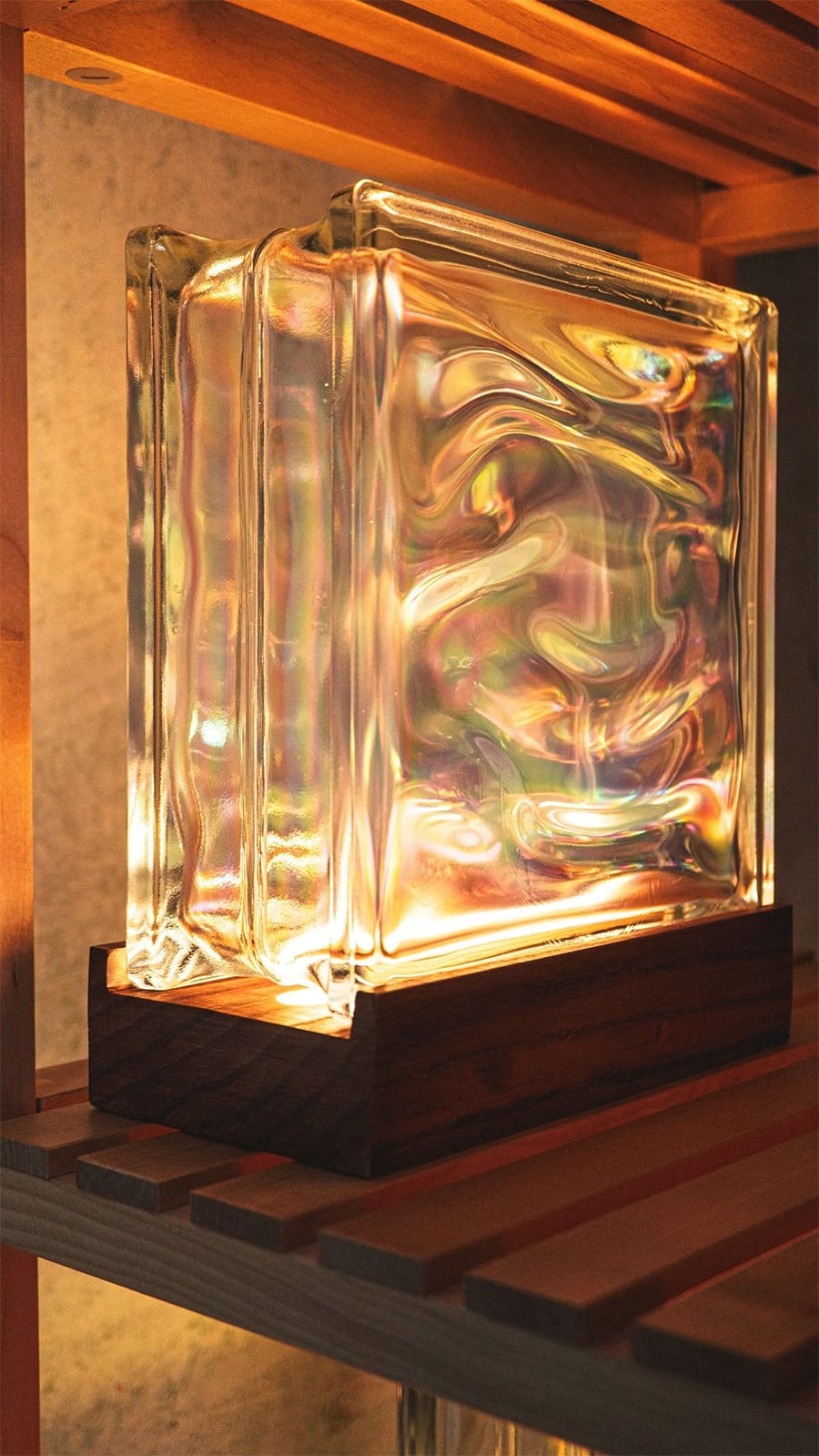 A glowing rectangular glass block with a wavy texture is placed upright on a wooden base, set on a slatted wooden shelf. Warm light shines through the glass, creating colorful reflections.