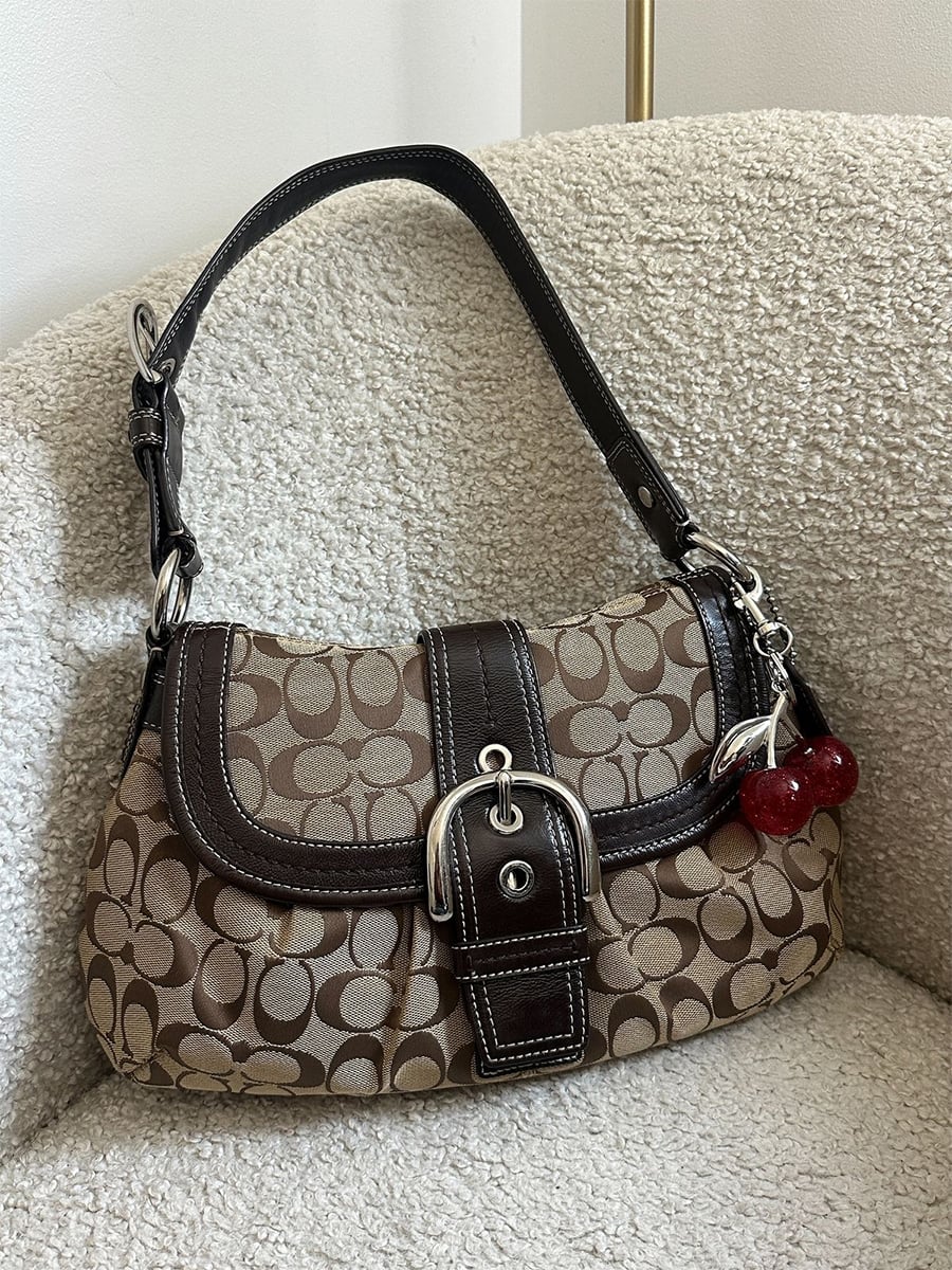 A brown Coach handbag with a signature "C" pattern, dark brown leather trim, silver buckle, and red cherry keychain, resting on a textured beige chair.