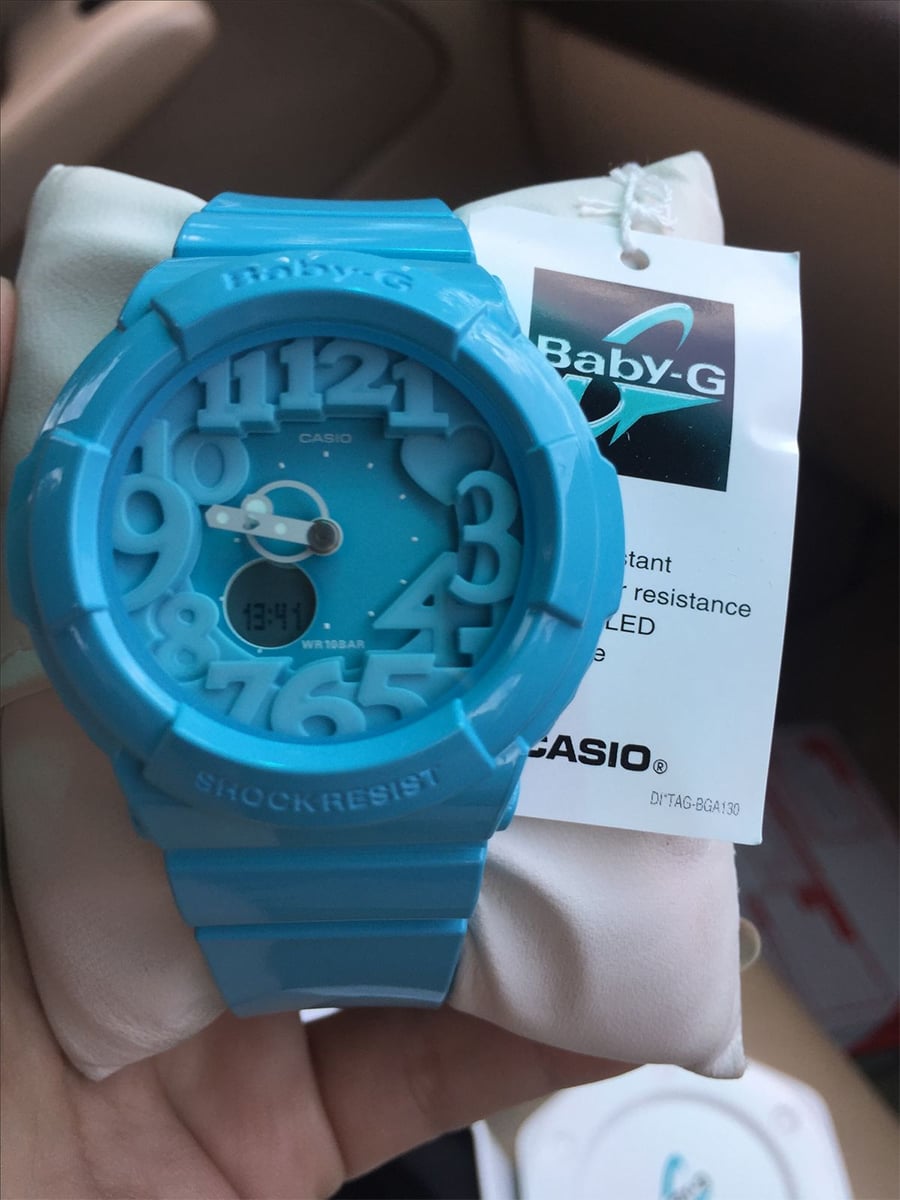 A hand holds a blue Casio Baby-G wristwatch with a round face, large numbers, and both analog and digital displays. The watch rests on a white cushion with its product tag attached.