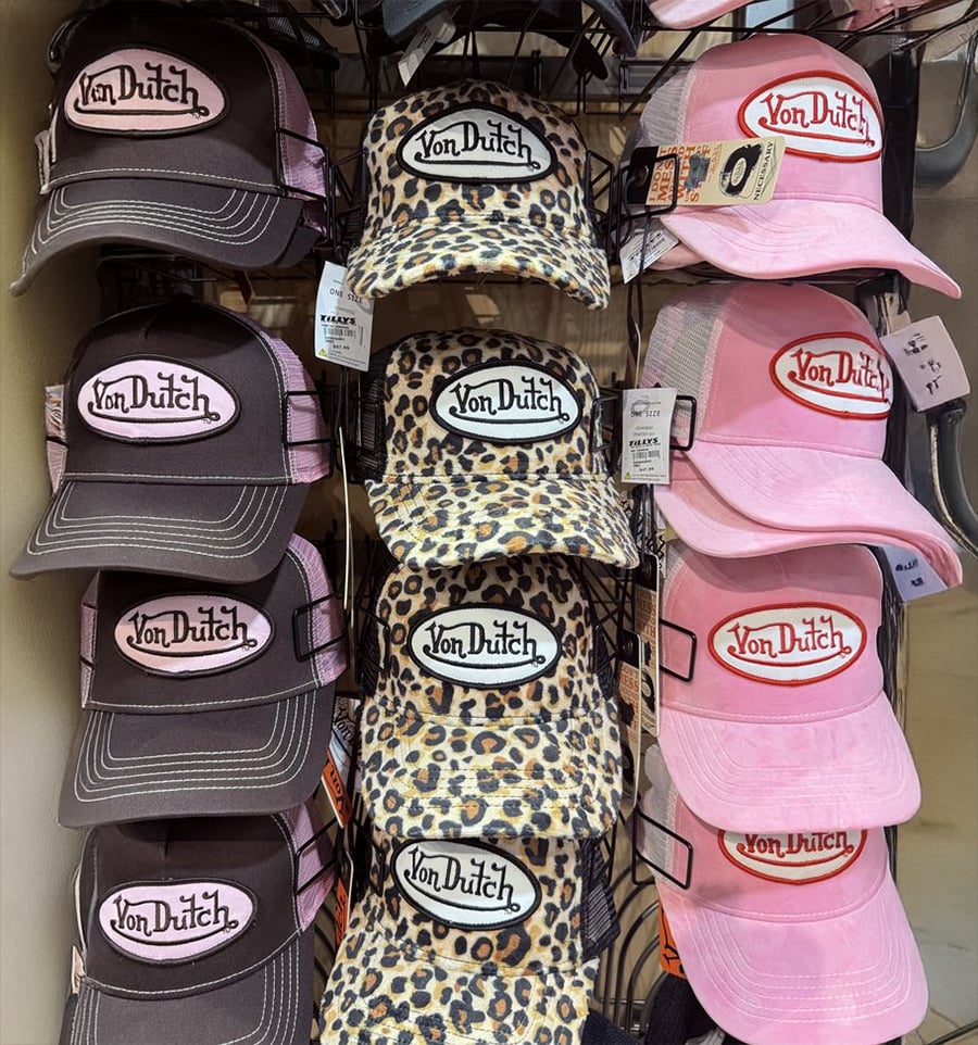 A display of Von Dutch hats in various colors and patterns, including pink, leopard print, and brown, arranged in three columns on a shop shelf. Each hat has a prominent Von Dutch logo patch.