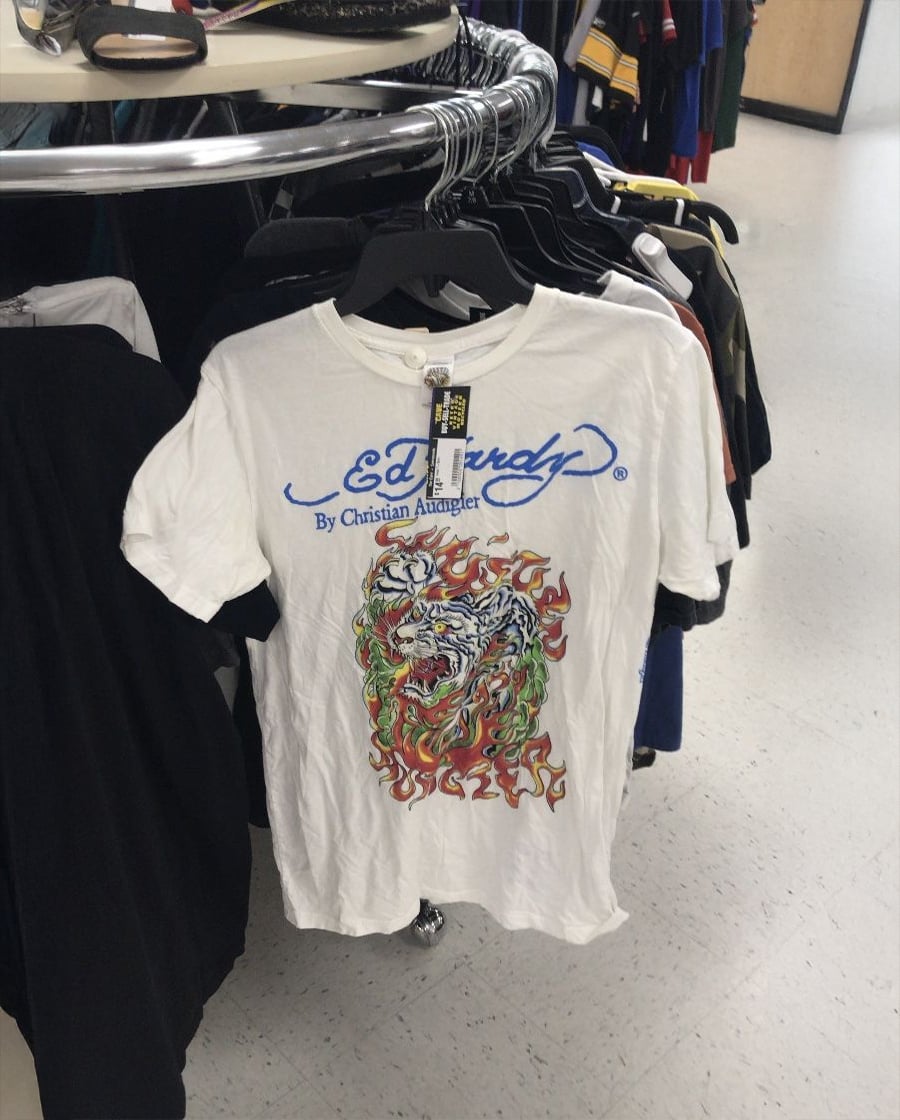 A white Ed Hardy T-shirt with a colorful tiger graphic and flames hangs on a black hanger on a clothing rack in a store.