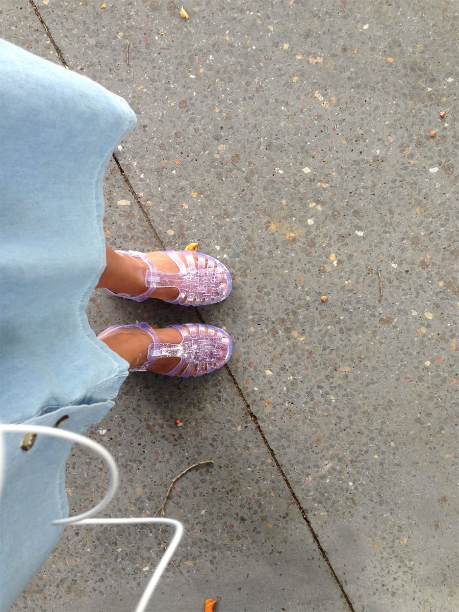 Person wearing a light blue skirt and sparkly purple jelly sandals stands on a speckled concrete surface. White earbud cables are visible in the lower left corner of the image.