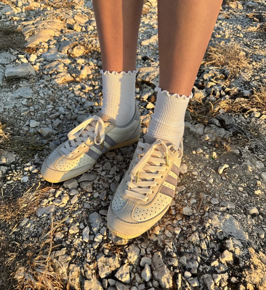 Person wearing white socks with ruffled edges and beige sneakers with gray and purple stripes, standing on rocky ground in warm sunlight.