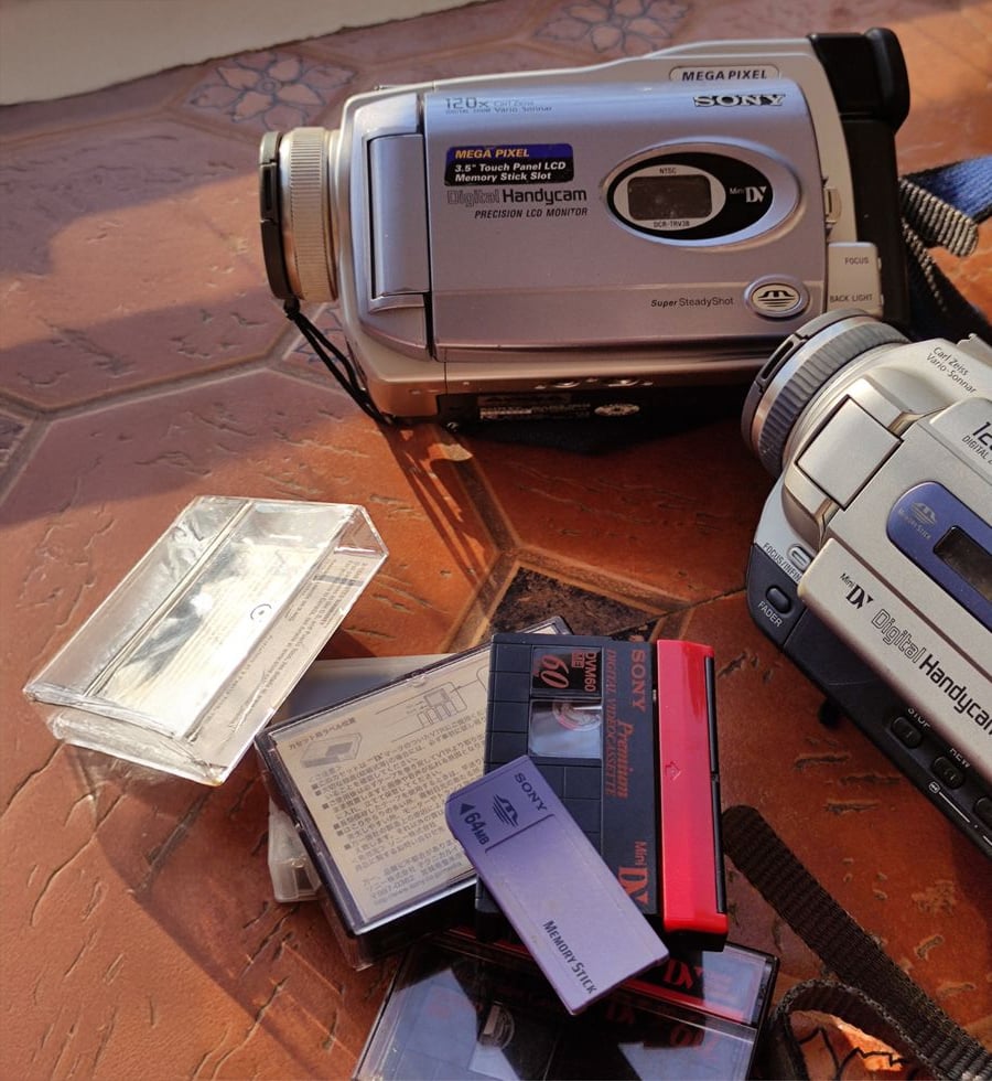 Two vintage Sony Handycams and several MiniDV tapes are on a textured surface under warm sunlight. The tapes, some labeled, are scattered near empty cases. Shadows are visible across the scene.