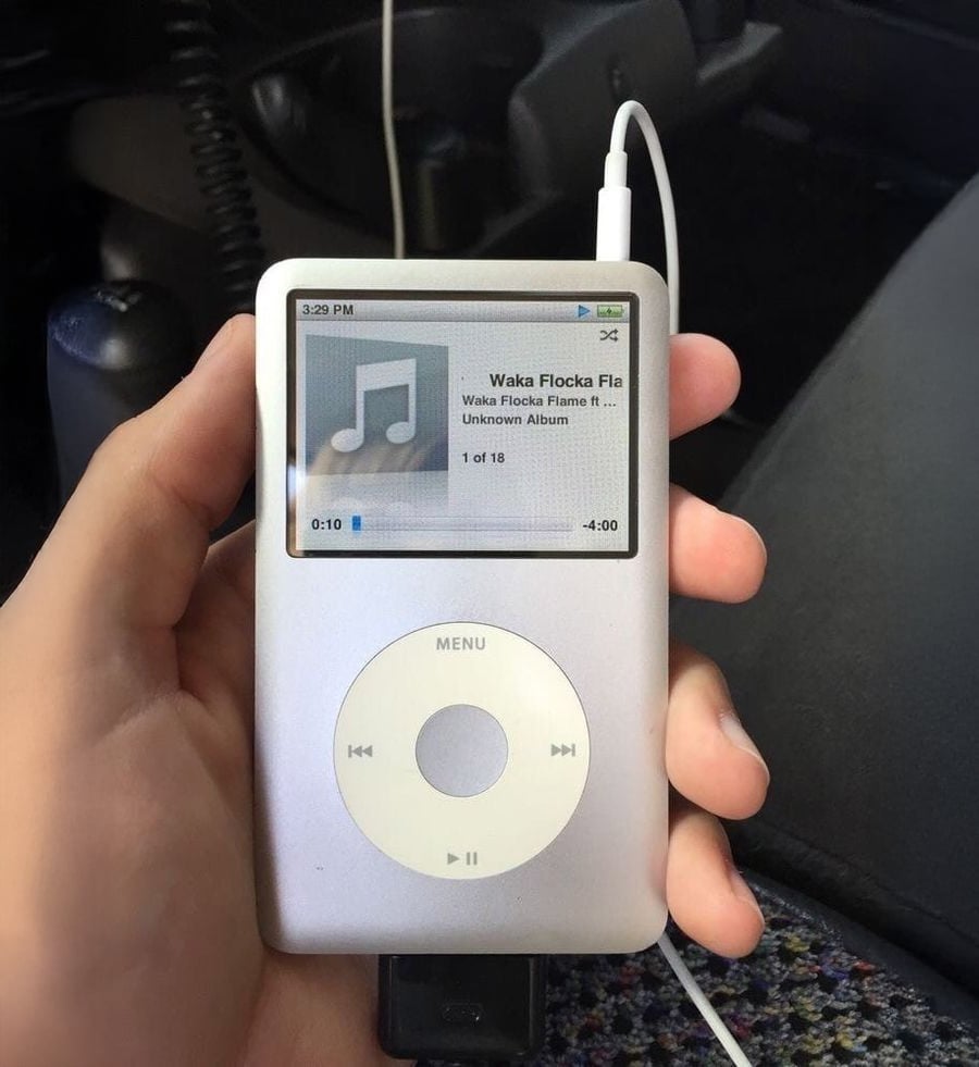 A hand holds a white iPod Classic displaying the song "Waka Flocka Flame" on its screen, with white earphones plugged in. The background shows part of a car interior.