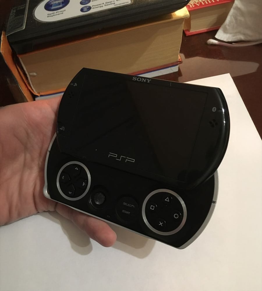 A hand holds a black Sony PSP Go gaming device with its screen slid open. Books and a white object are in the background on a brown surface.