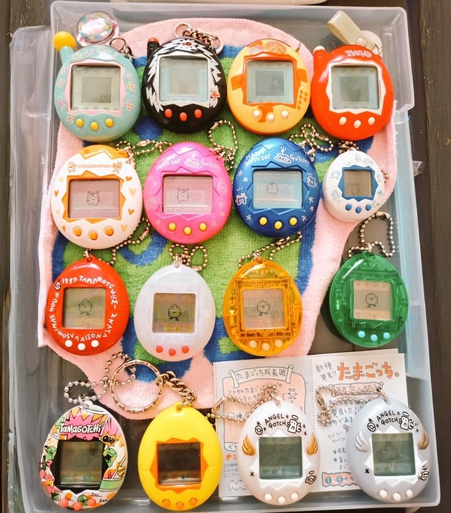A collection of colorful Tamagotchi virtual pets is neatly arranged in a box, with various shapes and designs, some themed as animals, all with keychains attached. Instructions and papers are included at the bottom.