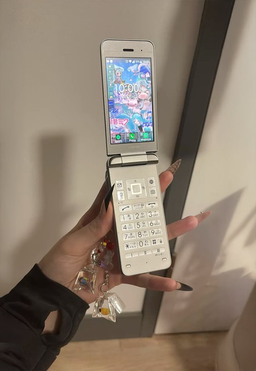 A hand with long nails holds an open silver flip phone decorated with colorful charms. The phone’s screen displays a bright anime-themed wallpaper and various app icons.