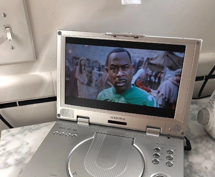 A portable DVD player on a marble counter displays a scene from a medieval-themed movie featuring a man in a green shirt looking concerned.