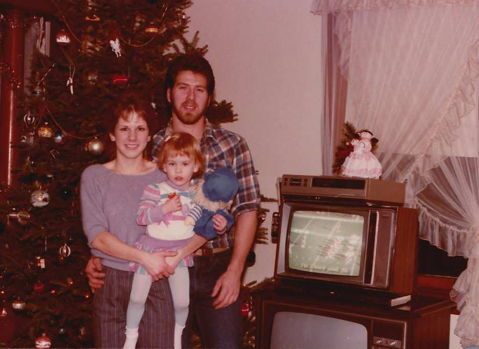 A family poses in front of a decorated Christmas tree. Two adults stand with a young child holding a doll. Next to them is a TV showing a football game with another smaller television stacked above it. Curtains cover a window behind them.