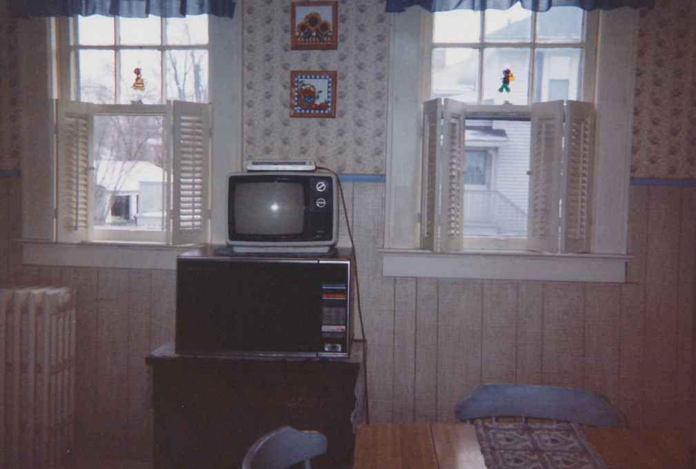 A vintage TV sits on top of a stereo system between two windows with half-open shutters in a cozy, old-fashioned room with floral wallpaper and wood paneling. A dining table and chairs are partially visible in the foreground.